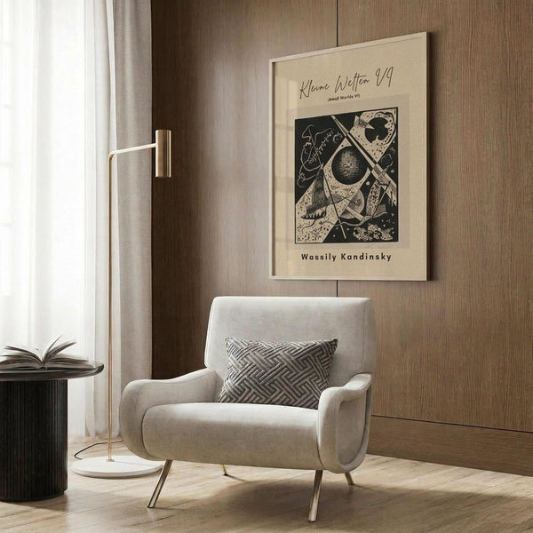 May include: Abstract geometric art poster by Wassily Kandinsky displayed in modern living room with warm wood paneling. Beige upholstered chair with geometric patterned pillow sits beside brass floor lamp and dark round side table with open book. Cream-colored poster features black and tan abstract shapes, circles, and lines in modernist style, mounted on wood wall near window with light curtains.
