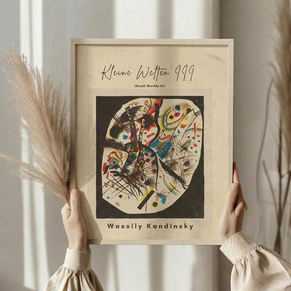May include: Person holding framed abstract art print featuring vibrant geometric shapes, lines and forms in red, blue, yellow and black within a circular composition. Light wood frame against cream matting. Dried pampas grass visible on left. Person wearing beige ruffled sleeve top in soft natural lighting by window with white curtains.