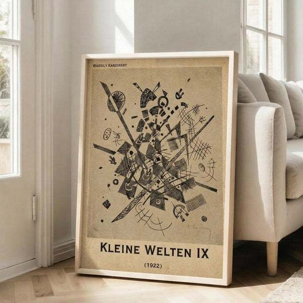 May include: Framed abstract geometric art print with black angular shapes, lines, and patterns on beige textured background, displayed in light wooden frame leaning against white wall corner next to beige upholstered chair in bright modern living room with large windows and natural sunlight streaming across hardwood floors.