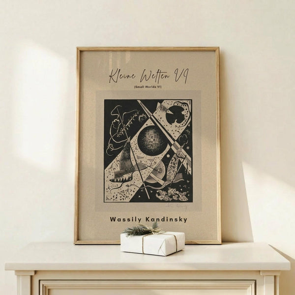 May include: Framed abstract art print by Wassily Kandinsky featuring geometric shapes, circles, and linear elements in black ink on cream paper, displayed on light wood surface beside small white gift box with twine and evergreen sprig against neutral wall