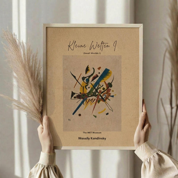 May include: Person in cream ruffled sleeves holding framed abstract art print with colorful geometric shapes, lines and forms in yellow, blue, red and green on beige background, alongside dried pampas grass stems against soft neutral curtained window setting
