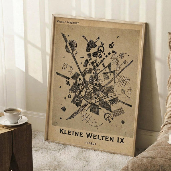 May include: Framed abstract art print with geometric black shapes, lines, and patterns on beige background, displayed in modern room setting next to coffee cup on wooden block and textured throw blanket on white fluffy rug.