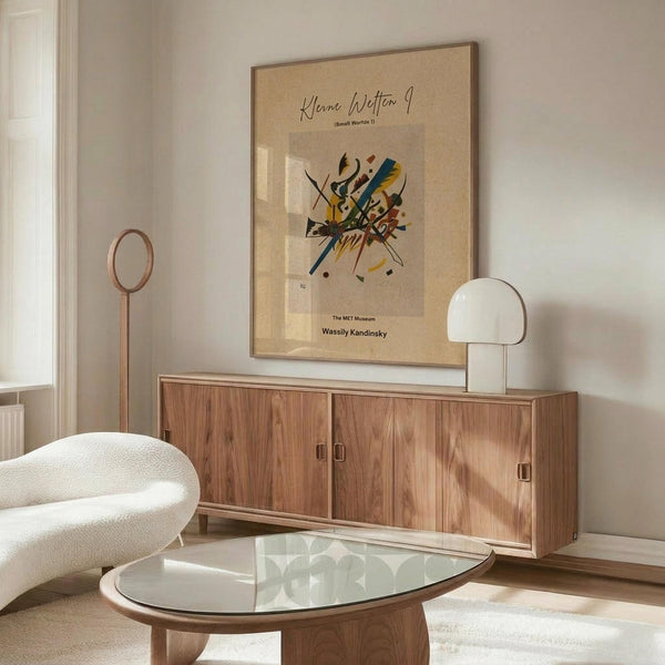 May include: Abstract colorful artwork poster with geometric shapes and lines in yellow, blue, red, and black, displayed in thin wood frame above mid-century modern walnut credenza. Modern living room setting features white curved chair, glass oval coffee table, mushroom table lamp, and brass floor lamp against neutral walls.