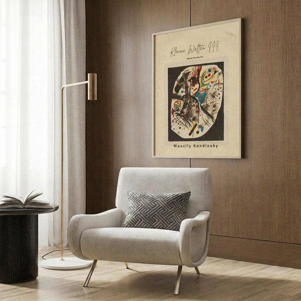 May include: Framed Wassily Kandinsky abstract art print with colorful geometric shapes in circular composition on cream background, displayed on wood paneled wall above modern beige armchair with geometric pillow, beside brass floor lamp and dark side table with open book