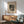 May include: Framed abstract art print with geometric shapes, circles, and linear elements in black and cream tones, displayed above a mid-century modern wooden credenza with sliding doors. Green upholstered armchair with wooden frame sits nearby, along with a small round side table and ceramic table lamp. Warm natural light creates shadows across the minimalist room setting with white walls.