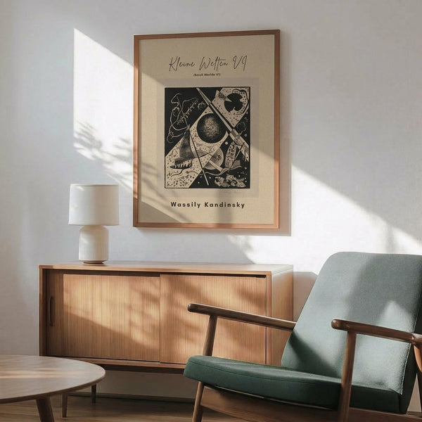 May include: Framed abstract art print with geometric shapes, circles, and linear elements in black and cream tones, displayed above a mid-century modern wooden credenza with sliding doors. Green upholstered armchair with wooden frame sits nearby, along with a small round side table and ceramic table lamp. Warm natural light creates shadows across the minimalist room setting with white walls.
