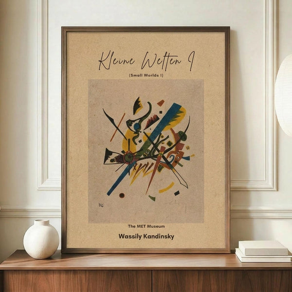 May include: Framed abstract art print with colorful geometric shapes and lines in yellow, blue, red, and green on beige background, displayed on wooden sideboard next to white ceramic vase and stack of books in modern interior with cream walls and white molding.