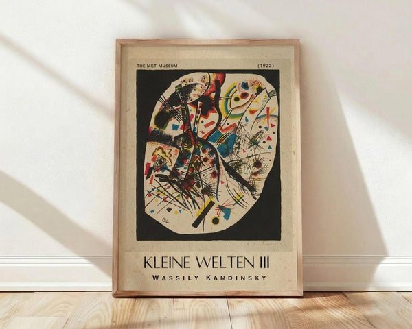 May include: Framed vintage museum poster featuring Wassily Kandinsky's abstract circular composition with bold geometric shapes, lines, and vibrant colors including red, blue, yellow, and black against cream background. Wooden frame rests on light hardwood floor against white wall with soft natural lighting creating diagonal shadows.