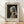 May include: Framed art print of Vermeer's Girl with a Pearl Earring displayed against white textured wall. Shows young woman in blue and yellow turban with luminous pearl earring against dark background. Natural wood frame with cream matting, sitting on light wooden floor with soft window shadows.