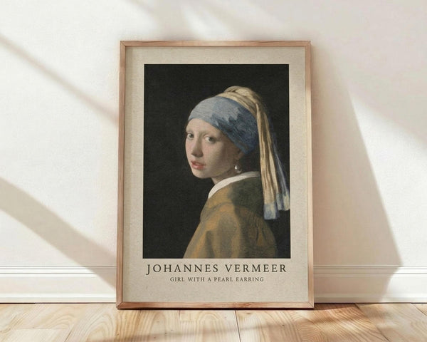 May include: Framed art print of Vermeer's Girl with a Pearl Earring displayed against white textured wall. Shows young woman in blue and yellow turban with luminous pearl earring against dark background. Natural wood frame with cream matting, sitting on light wooden floor with soft window shadows.