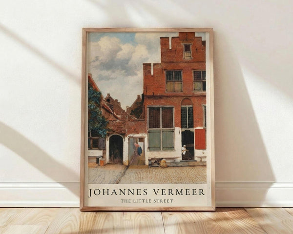 May include: Framed art print of Johannes Vermeer's classic Dutch painting showing a cobblestone courtyard with red brick houses, small figures in period dress, and cloudy sky. The wooden-framed poster leans against white walls on light wood flooring, with natural window light creating soft shadows across the scene.