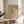 May include: Framed beige poster with abstract black line drawings featuring geometric shapes, dots, and angular forms displayed on a round wooden side table next to a light gray upholstered chair. Natural sunlight creates soft shadows across the minimalist white wall setting, with green plant leaves visible in the corner.