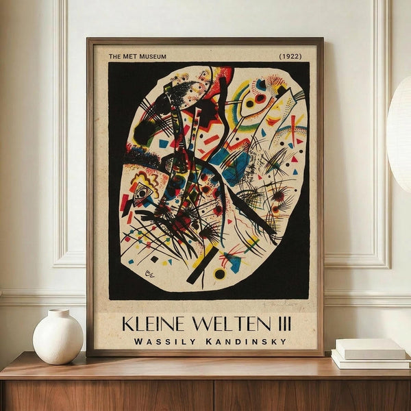 May include: Framed abstract art print displayed on wooden sideboard featuring circular composition with geometric shapes, lines, and colorful forms in Kandinsky's distinctive style. Black background with vibrant reds, blues, yellows. Museum exhibition poster format with beige border. Accompanied by white ceramic vase and stacked books in modern interior setting with white paneled walls.