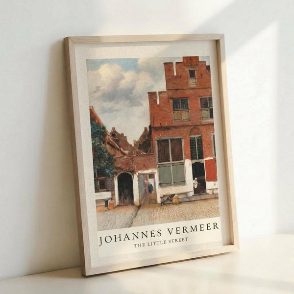 May include: Framed art print displaying a Dutch Golden Age street scene with red brick buildings, cobblestone courtyard, and cloudy sky. The reproduction shows detailed architecture including a distinctive stepped-gable house and multi-story brick structure. Light wooden frame against white wall, positioned at an angle showing depth and shadow.