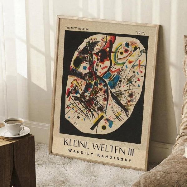 May include: Abstract Kandinsky poster in light wood frame leaning against white wall in cozy room setting. The artwork features colorful geometric shapes, lines, and forms in red, blue, yellow, and black arranged in an oval composition. Small wooden side table with white coffee cup and saucer sits nearby on textured cream rug. Soft natural lighting creates gentle shadows on the wall.