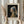 May include: Person in cream blouse holding framed art print of Vermeer's Girl with a Pearl Earring in light wood frame, alongside dried wheat stalks, against white curtained window background. Print shows woman in blue turban with pearl earring on dark background, with artist name and title text below image.