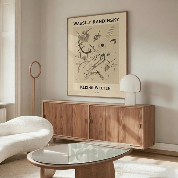 May include: Framed Kandinsky abstract art print displayed above wooden sideboard in minimalist living room. Black line drawings on cream background show geometric shapes, dots, and angular forms. White modern chair and glass coffee table visible. Warm wood tones throughout mid-century modern interior with natural lighting.
