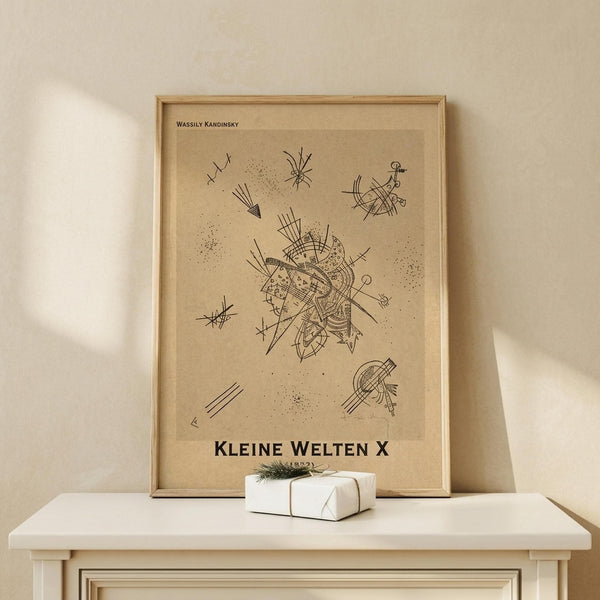 May include: Abstract art print in light wood frame showing geometric black line drawings on beige background, featuring intersecting lines, dots, and angular shapes in Kandinsky's distinctive style. Displayed on white wooden table beside small wrapped white gift box with twine and greenery sprig against cream colored wall with soft natural lighting.