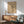 May include: Framed Kandinsky abstract art print with geometric lines and shapes on beige background, displayed above wooden sideboard with white ceramic lamp. Mid-century modern sage green armchair with wooden arms sits nearby. Warm sunlight creates shadows across white wall and wooden furniture in minimalist living room setting.
