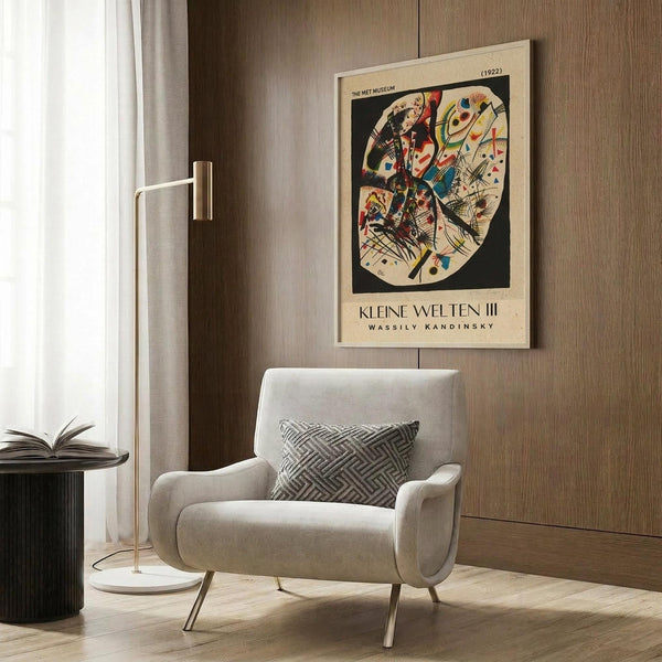 May include: Framed vintage art poster featuring Wassily Kandinsky's abstract circular composition with vibrant geometric shapes, lines, and forms in red, blue, yellow, and black. Displayed on wood-paneled wall above modern beige armchair with geometric patterned pillow. Room includes brass floor lamp and side table with open book.