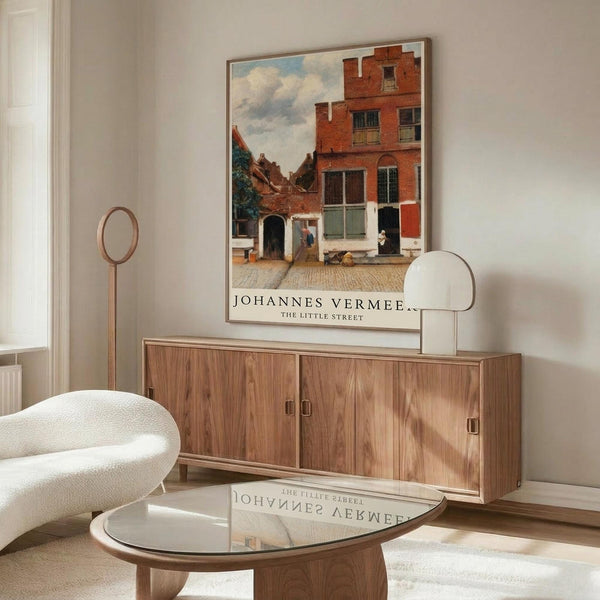 May include: A framed art print featuring Vermeer's painting of Dutch buildings with red brick facades and a cobblestone courtyard under a cloudy sky. The poster hangs above a mid-century modern walnut credenza in a minimalist living room with white walls, accompanied by a cream upholstered chair, brass floor lamp, glass coffee table, and white table lamp.