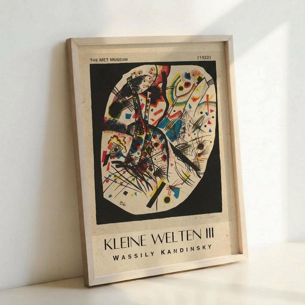 May include: Framed poster print of Wassily Kandinsky's abstract artwork featuring circular composition with vibrant geometric shapes, lines, and forms in red, blue, yellow, and black colors against cream background. Text reads "The Met Museum (1922)" at top and "Kleine Welten III Wassily Kandinsky" at bottom. Light wood frame displayed against white wall, leaning on surface with soft natural lighting creating subtle shadows.