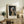 May include: Framed art print of Vermeer's Girl with a Pearl Earring displayed on rustic wooden sideboard in cozy living room setting. Print shows young woman in blue and yellow headwrap with pearl earring against dark background. Warm neutral decor with fern plant, books, ceramic vase, and beige armchair visible.
