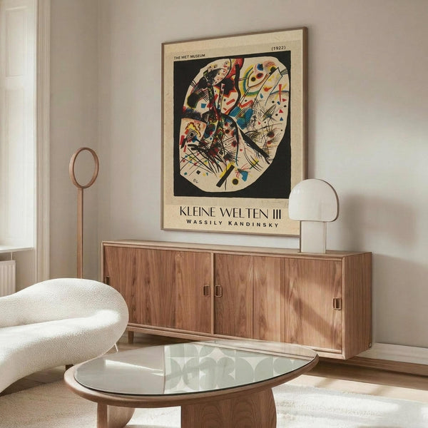 May include: Kandinsky abstract art poster displayed above wooden sideboard in modern living room. Colorful geometric shapes and lines within oval form on cream background. White curved chair, glass coffee table, and brass floor lamp visible. Poster shows vibrant reds, blues, yellows against black background with museum attribution text below artwork.