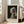 May include: Framed art print of Vermeer's Girl with a Pearl Earring displayed in bright living room with beige sofa. Classic portrait shows young woman in blue and yellow turban with pearl earring against dark background. Light wood frame with white mat, positioned on wooden floor near large windows with natural sunlight streaming in.