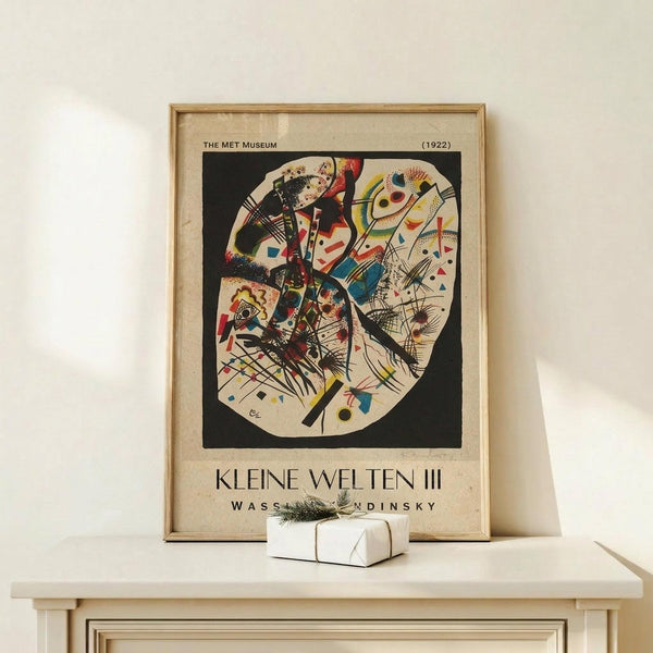 May include: Framed museum poster featuring Kandinsky's abstract circular composition with bold geometric shapes, lines, and vibrant colors including red, blue, yellow, and black on cream background. Wooden frame sits on light wooden surface next to small wrapped gift with twine and greenery sprig against neutral wall.