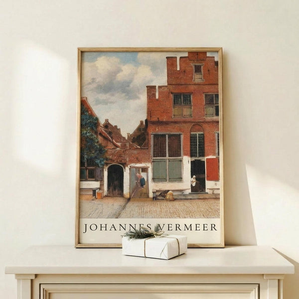 May include: Framed art print of a Dutch Golden Age street scene with red brick buildings, cobblestone courtyard, and small figures in period dress under a cloudy sky. The poster sits on a light wood shelf against a cream wall, with a small white wrapped gift with greenery beside it. Natural lighting creates soft shadows across the minimalist display.
