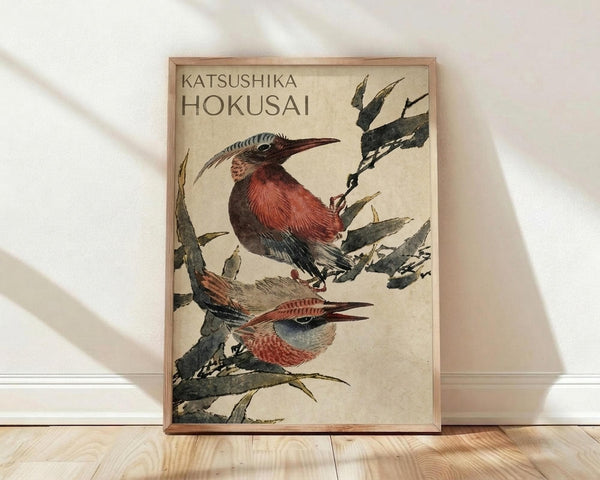 May include: Framed vintage Japanese woodblock print featuring two colorful birds perched on bamboo branches against a cream background. The artwork shows red-breasted birds with long beaks in traditional ukiyo-e style with green foliage. The wooden frame leans against a white wall on light wood flooring with soft natural lighting casting shadows.