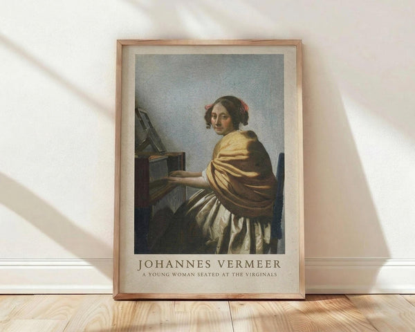 May include: Framed art print of Vermeer's classical painting showing a young woman in golden yellow and white dress seated at virginals, looking over her shoulder. Natural wood frame with cream matting, displayed on light wooden floor against white wall with soft natural lighting creating diagonal shadows.
