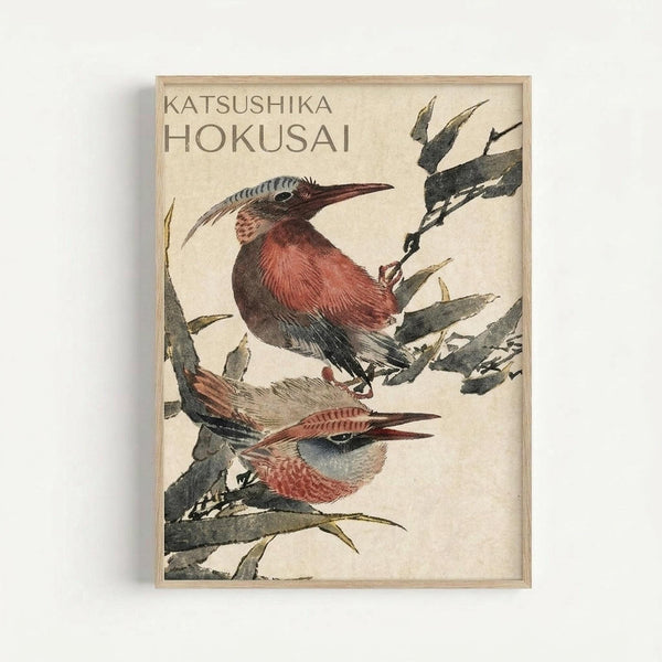 Japanese woodblock print style artwork featuring two birds with reddish-brown and blue-gray plumage perched on bamboo or reed stems. The birds have long, curved beaks and detailed feather patterns rendered in traditional ukiyo-e style with muted earth tones against a cream background. Displayed in a light wooden frame on a white wall.