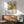 May include: A framed Japanese art print featuring white hydrangea flowers and a black bird on yellow background, displayed on white wall above wooden sideboard. Mid-century modern mint green armchair with wooden arms sits nearby, along with white ceramic table lamp and round wooden coffee table. Natural sunlight creates geometric shadows across the minimalist room setting.