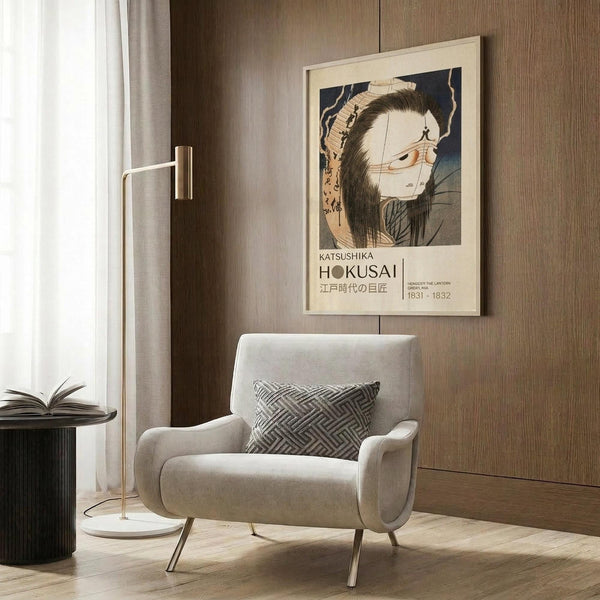 May include: Framed poster featuring Japanese ukiyo-e artwork of a ghostly figure with long dark hair and pale face against dark background, displayed above beige upholstered armchair with geometric patterned pillow in modern living room setting with wood paneled walls, brass floor lamp, and dark round side table with open book