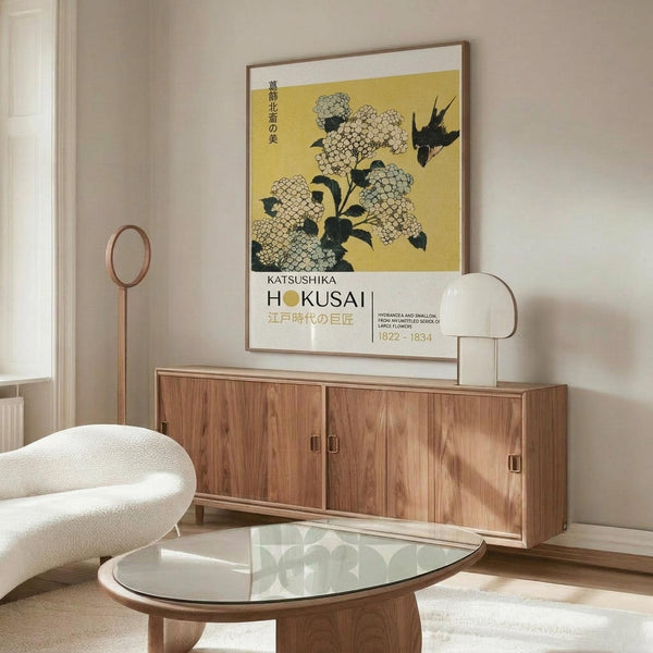 May include: Japanese art poster featuring white hydrangea flowers and black bird on yellow background, displayed in thin brown frame above wooden sideboard. Modern living room setting with cream chair, glass oval coffee table, and floor lamp. Clean minimalist interior with neutral walls and natural lighting.