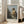 May include: Framed art print of Vermeer's classical painting showing a young woman in golden yellow and white robes seated at a virginal keyboard instrument, displayed in a bright modern living room with beige sofa, white walls, and large windows with natural sunlight streaming across wooden floors.
