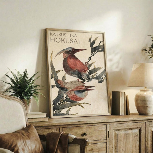 May include: Framed vintage-style poster featuring two detailed birds with reddish-brown and gray plumage perched on leafy branches, displayed on a rustic wooden sideboard alongside a fern plant, ceramic vase, and books in a neutral-toned living room setting with beige chair visible.