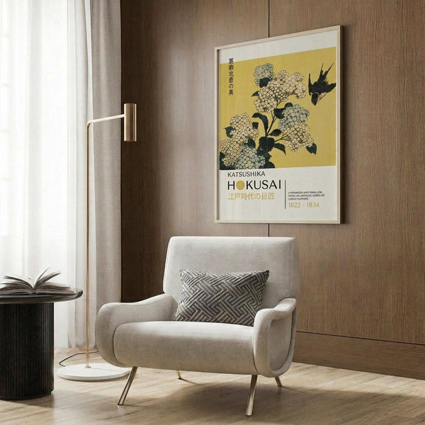 May include: A framed Japanese art poster featuring hydrangea flowers and a black bird on yellow background, displayed on wood-paneled wall above a beige upholstered armchair with geometric patterned pillow. Modern reading corner with brass floor lamp, dark round side table with open book, and white curtains visible.