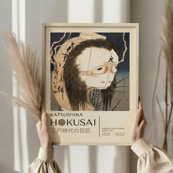 May include: Hands holding a framed Japanese woodblock print featuring a ghostly female figure with long dark hair and pale skin against a blue background. The artwork shows traditional ukiyo-e style with Japanese text. Person wears cream-colored clothing with ruffled sleeves, dried pampas grass visible to the left, neutral beige wall background.