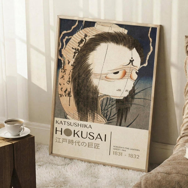 May include: A framed vintage Japanese woodblock print featuring a pale ghostly figure with long black hair and traditional dress against a dark blue background with Japanese text. The poster is displayed in a light wooden frame, leaning against white walls in a cozy room setting with a coffee cup on a wooden surface and soft textured blankets nearby.