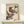 May include: Framed vintage Japanese woodblock print featuring two colorful birds with reddish-pink breasts and long curved beaks perched on dark green bamboo or grass stalks against cream background. Print displayed on light wooden surface next to small white wrapped gift with evergreen sprig. Natural lighting creates soft shadows on neutral wall.