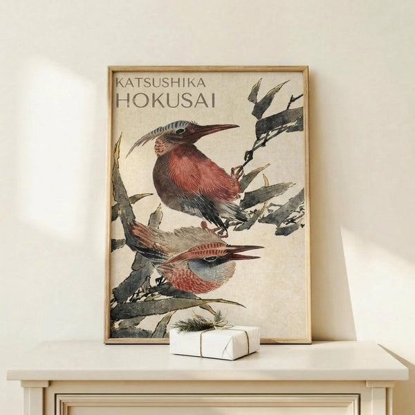 May include: Framed vintage Japanese woodblock print featuring two colorful birds with reddish-pink breasts and long curved beaks perched on dark green bamboo or grass stalks against cream background. Print displayed on light wooden surface next to small white wrapped gift with evergreen sprig. Natural lighting creates soft shadows on neutral wall.