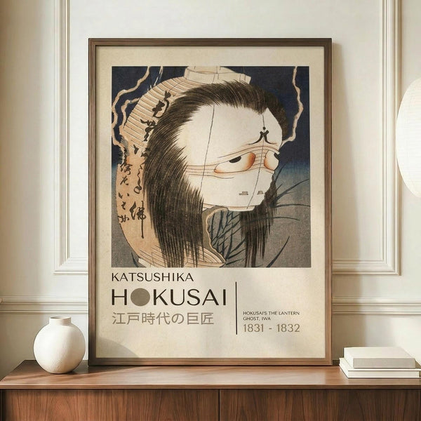 May include: A framed Japanese woodblock print poster featuring a ghostly female figure with long black hair and pale white face, holding a paper lantern. The artwork shows traditional ukiyo-e style with muted colors against a dark blue background. Text below reads "Katsushika Hokusai" with Japanese characters and dates "1831-1832". The wooden-framed print sits on a light wood sideboard next to a white ceramic vase and books, displayed against cream colored wainscoting walls.