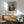 May include: Framed art print of Johannes Vermeer's classical painting showing a young woman in golden yellow and white period dress seated at a virginals keyboard instrument. The poster features warm earth tones with museum-style text below the image. Displayed on white wall above wooden mid-century modern credenza with ceramic table lamp, next to sage green upholstered armchair with wooden frame. Natural sunlight creates diagonal shadows across the minimalist interior scene.