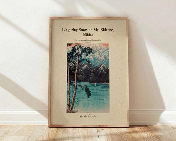 May include: Framed vintage Japanese woodblock print showing a winter mountain landscape with snow-capped peaks, a lone pine tree in foreground, and serene blue lake reflecting the scenery. The artwork features traditional ukiyo-e style with muted blues, greens, and coral sky tones on cream matting in natural wood frame, displayed on light wooden floor against white wall with soft natural lighting.