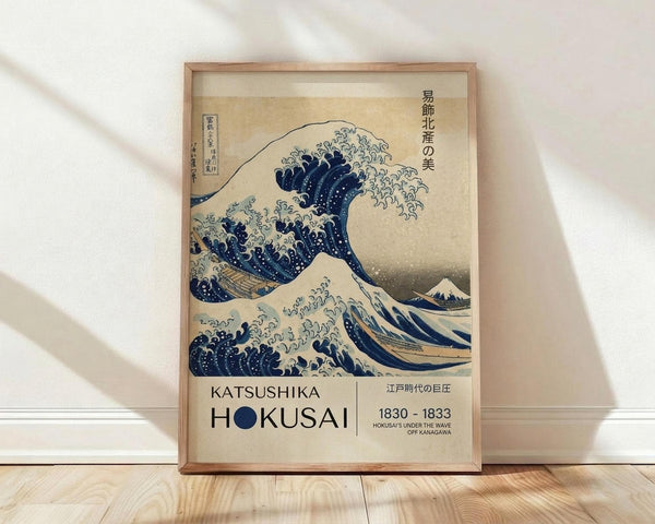 May include: Framed reproduction of Hokusai's famous Great Wave woodblock print displayed against white wall on wooden floor. The artwork shows a large blue wave with white foam threatening boats, with Mount Fuji visible in background. Poster features exhibition-style text reading "Katsushika Hokusai 1830-1833" and Japanese characters on cream-colored paper in simple wooden frame.