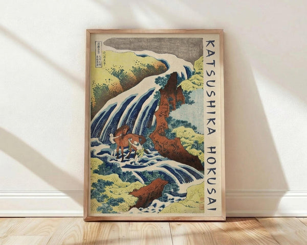 May include: Framed Japanese woodblock print artwork showing a dramatic waterfall cascading over brown rocky cliffs with white foam and blue water streams. Two small figures visible near the base of the falls. Artwork features traditional ukiyo-e style with yellowy-green foliage, gray sky, and Japanese text characters along the right side. Light wood frame displayed against white wall on wooden floor with natural lighting.