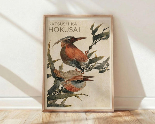 May include: Framed Japanese-style artwork showing two birds with russet-orange breasts and long curved beaks perched on leafy branches, displayed in a light wood frame against a white textured wall on natural wood flooring with soft natural lighting creating diagonal shadows.